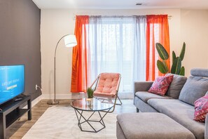 Bright living room with big windows, plush grey sofa adorned with colorful throw pillows, and a cozy pink accent chair. #LuxuriousLiving #CozyRetreat
