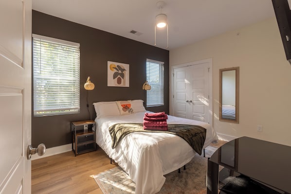 In the photo, a cozy bedroom exudes tranquility with its soft pastel color palette. A queen-sized bed with a tufted headboard is adorned with plush pillows and a fluffy duvet, inviting relaxation. Two wooden nightstands flank the bed, each topped with a minimalist lamp and a small potted plant. Sheer curtains drape over large windows, allowing diffused sunlight to fill the room...