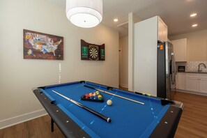 The photo features a lively game room designed for entertainment and leisure. At the center stands a classic Pool table with a rich walnut finish and a deep blue felt surface, illuminated by a trio of hanging pendant lights above. To one side, a sleek Dartboard  flashes colorful graphics, offering a touch of nostalgia. 