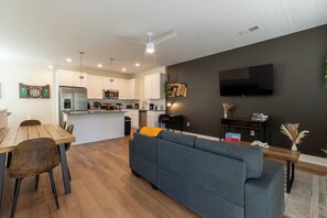 The photo depicts a spacious living room that combines comfort and elegance. A large sectional sofa upholstered in gray fabric faces a modern fireplace framed by a sleek mantel. Above the fireplace hangs an abstract painting that adds a splash of color to the neutral tones of the room. Floor-to-ceiling windows offer a panoramic view of the city skyline, allowing natural light t...