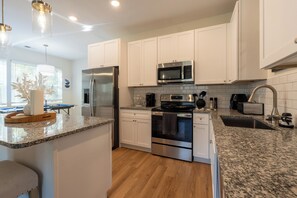 The photo showcases a modern kitchen bathed in natural light pouring through a large window above the sink. Sleek white cabinets line the walls, contrasted by a dark granite countertop that adds depth to the space. Stainless steel appliances—including a refrigerator, oven, and dishwasher—reflect the clean lines and minimalist design of the room. A central island features bar st...