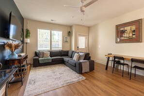 The photo depicts a spacious living room that combines comfort and elegance. A large sectional sofa upholstered in gray fabric faces a modern fireplace framed by a sleek mantel. Above the fireplace hangs an abstract painting that adds a splash of color to the neutral tones of the room. Floor-to-ceiling windows offer a panoramic view of the city skyline, allowing natural light t...