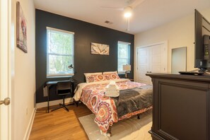 In the photo, a cozy bedroom exudes tranquility with its soft pastel color palette. A queen-sized bed with a tufted headboard is adorned with plush pillows and a fluffy duvet, inviting relaxation. Two wooden nightstands flank the bed, each topped with a minimalist lamp and a small potted plant. Sheer curtains drape over large windows, allowing diffused sunlight to fill the room...
