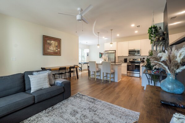 The photo depicts a spacious living room that combines comfort and elegance. A large sectional sofa upholstered in gray fabric faces a modern fireplace framed by a sleek mantel. Above the fireplace hangs an abstract painting that adds a splash of color to the neutral tones of the room. Floor-to-ceiling windows offer a panoramic view of the city skyline, allowing natural light t...