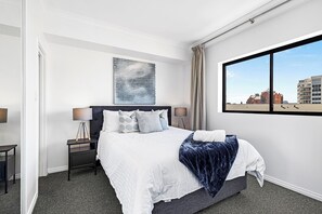Unwind in a serene bedroom featuring a plush king-size bed with crisp white linens, a dark blue throw, and a city view from the window.