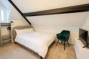 Charming attic bedroom with exposed beam and plush bedding, complete with TV, velvet green armchair and open wardrobe. The sloped ceiling and soft lighting create a warm, inviting atmosphere. (1)