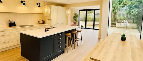 Spacious open-concept kitchen and dining area with natural light streaming through large glass windows. Features a sleek navy island, cozy wood tones, and a view of a green garden—perfect for family gatherings and entertaining!