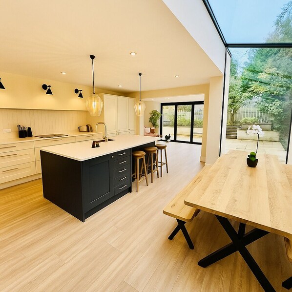 Spacious open-concept kitchen and dining area with natural light streaming through large glass windows. Features a sleek navy island, cozy wood tones, and a view of a green garden—perfect for family gatherings and entertaining!
