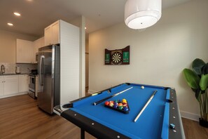 The photo features a lively game room designed for entertainment and leisure. At the center stands a classic Pool table with a rich walnut finish and a deep blue felt surface, illuminated by a trio of hanging pendant lights above. To one side, a sleek Dartboard  flashes colorful graphics, offering a touch of nostalgia. 
