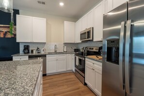 The photo showcases a modern kitchen bathed in natural light pouring through a large window above the sink. Sleek white cabinets line the walls, contrasted by a dark granite countertop that adds depth to the space. Stainless steel appliances—including a refrigerator, oven, and dishwasher—reflect the clean lines and minimalist design of the room. A central island features bar st...