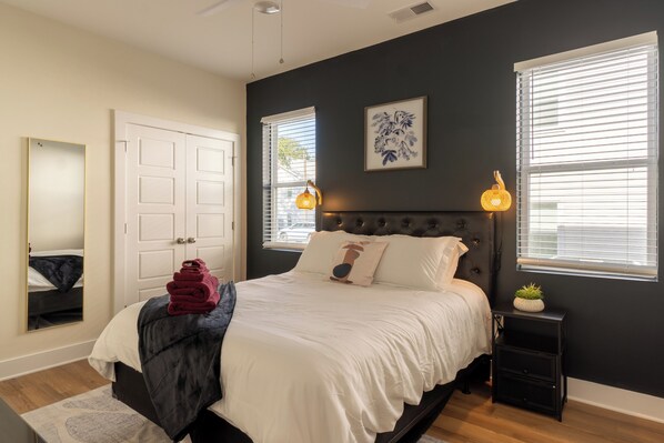 In the photo, a cozy bedroom exudes tranquility with its soft pastel color palette. A queen-sized bed with a tufted headboard is adorned with plush pillows and a fluffy duvet, inviting relaxation. Two wooden nightstands flank the bed, each topped with a minimalist lamp and a small potted plant. Sheer curtains drape over large windows, allowing diffused sunlight to fill the room...