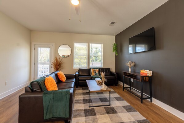 The photo depicts a spacious living room that combines comfort and elegance. A large sectional sofa upholstered in gray fabric faces a modern fireplace framed by a sleek mantel. Above the fireplace hangs an abstract painting that adds a splash of color to the neutral tones of the room. Floor-to-ceiling windows offer a panoramic view of the city skyline, allowing natural light t...