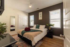 In the photo, a cozy bedroom exudes tranquility with its soft pastel color palette. A queen-sized bed with a tufted headboard is adorned with plush pillows and a fluffy duvet, inviting relaxation. Two wooden nightstands flank the bed, each topped with a minimalist lamp and a small potted plant. Sheer curtains drape over large windows, allowing diffused sunlight to fill the room...