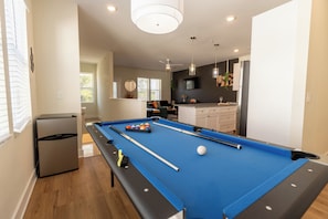 The photo features a lively game room designed for entertainment and leisure. At the center stands a classic Pool table with a rich walnut finish and a deep blue felt surface, illuminated by a trio of hanging pendant lights above. To one side, a sleek Dartboard  flashes colorful graphics, offering a touch of nostalgia. 