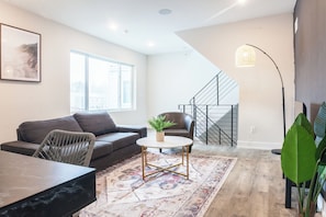 This serene living room features a calming grey color palette with a plush grey sofa, a sleek grey chair, and a modern coffee table. Lush greenery adds a touch of life, while a big window brings in natural light. Unwind by the TV or enjoy the city views in this tranquil space.