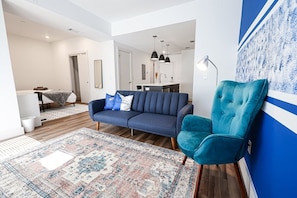 A modern living room features a navy blue sofa adorned with white pillows, complemented by a teal armchair. A multicolored rug anchors the space. The white and blue walls are decorated with abstract art, creating a vibrant yet cozy atmosphere #ColourfulInterior