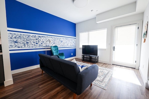 Our living room features a bold blue wall with a white and blue patterned stripe, complemented by contemporary furniture including a dark sofa, teal armchair, and a TV on a black stand; the room is illuminated by natural light from large windows. #SunshineVibes