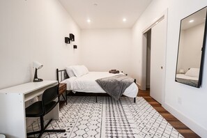 Experience the comfort and simplicity of this modern bedroom, featuring a cozy double bed. A functional workspace is provided by a sleek white desk paired with a comfortable black chair, ideal for business travelers or creative souls. A large mirror enhances the spacious feel, while the patterned rug adds a touch of elegance. 