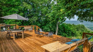 Spacious wooden deck overlooking a panoramic mountain landscape, with clear skies and lush greenery in the distance. Ideal for relaxing or entertaining while enjoying the natural beauty of the surroundings.
