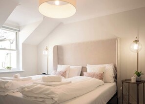 A serene bedroom bathed in natural light features a soft upholstered headboard, matching bedside lamps, and delicate pink cushions for a touch of charm.