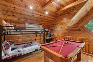 The open loft features a twin-over-full bunk bed with cozy patchwork bedding, rustic wood walls, and easy access to the arcade game and pool table—perfect for kids or fun-loving adults.