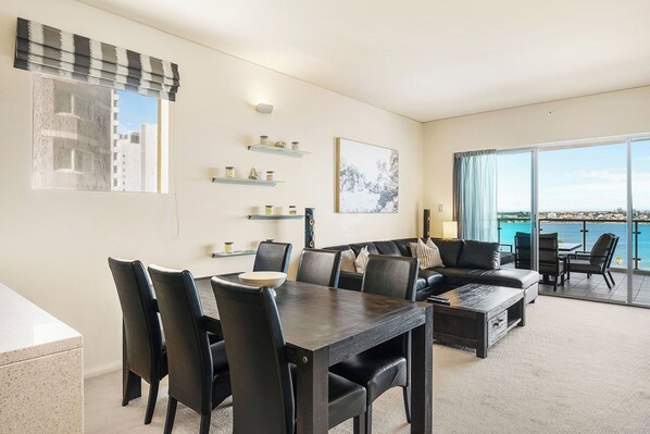 A bright and inviting open-plan living space, seamlessly blending a dining area and living room, with stunning waterfront views visible through the sliding glass doors. The space is designed for comfort and socializing, perfect for guests who appreciate modern amenities and scenic surroundings.