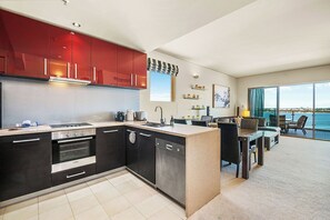A spacious and interconnected living space.  A glimpse of the living room with a plush sofa suggests a relaxing space for socializing. Features striking red glossy upper cabinets and dark wood lower cabinets, adding a vibrant and contemporary touch.