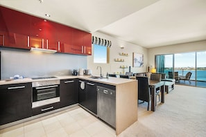 A spacious and interconnected living space. A glimpse of the living room with a plush sofa suggests a relaxing space for socializing. Features striking red glossy upper cabinets and dark wood lower cabinets, adding a vibrant and contemporary touch.