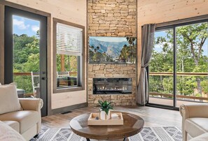 Living area is well decorated with electric firepace, Smart TV, comfy seating, and forest views.