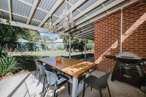 Step onto the covered patio, where a generous dining table and BBQ invite you to enjoy alfresco meals beneath glowing chandeliers, all set against the backdrop of a lush garden.