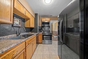 A cozy kitchen with wooden cabinets, granite countertops, a black refrigerator, and stainless steel appliances, set on a tiled floor and neutral walls.