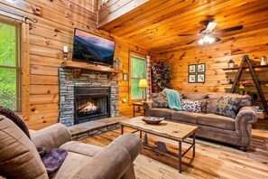 Relax in the inviting living room featuring a stone fireplace, plush seating, and rustic cabin charm, ideal for cozy nights and movie marathons.