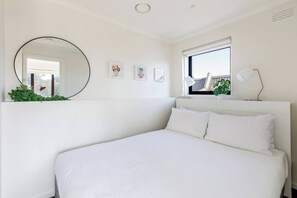Retreat to the queen-sized bed dressed in crisp white linen, with soft lighting and a blackout blind setting the scene for restful sleep.