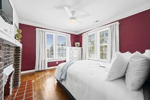 Welcome to our charming bungalow! The bedroom boasts rich red walls and a cozy white bed adorned with plush pillows and a light blue throw, adding a touch of elegance. A striking brick fireplace serves as the focal point, complemented by white linens