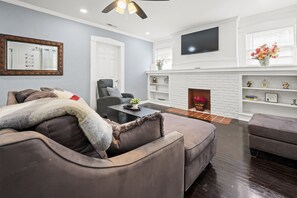 Welcome to our inviting living room, the space boasts a comfortable grey sectional sofa and a massage armchair, centered around a charming white brick fireplace. 