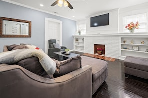 Welcome to our inviting living room, the space boasts a comfortable grey sectional sofa and a massage armchair, centered around a charming white brick fireplace.