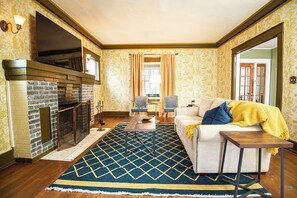 Cozy living room with vintage charm featuring a brick fireplace, floral wallpaper, and hardwood floors. A beige sofa, navy rug, and gold accents add warmth, while large windows and French doors bring in natural light for a welcoming atmosphere.