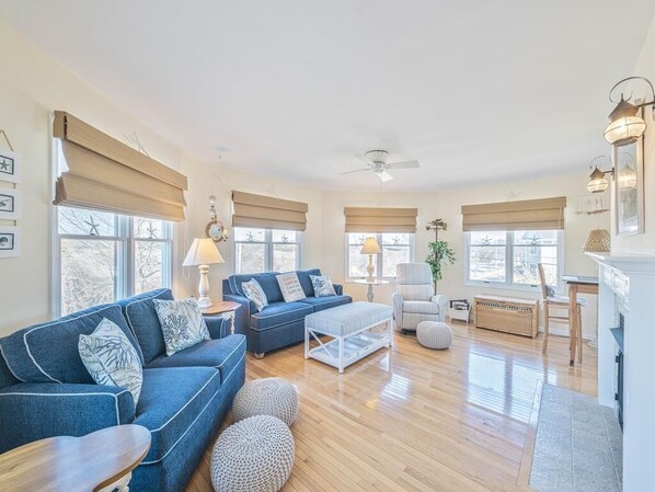 Relax in the spacious living area adorned with coastal decor, featuring a plush blue sofa and abundant natural light from large windows, perfect for family gatherings after a sun-soaked day at the beach.
