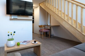 The living area includes a mounted TV and a dining table, highlighted by a staircase leading to additional space.