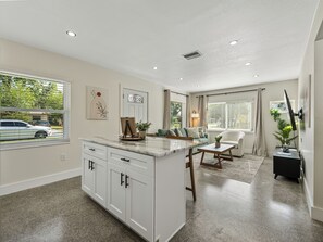 This modern living room features earth-toned decor, large windows for natural light, and cozy seating, including a swivel chair. A 65" TV is perfect for movie nights, while natural materials and soft lighting create a serene, inviting atmosphere. The space flows into the kitchen.