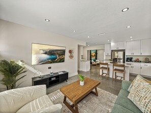 This modern living room features earth-toned decor, large windows for natural light, and cozy seating, including a swivel chair. A 65" TV is perfect for movie nights, while natural materials and soft lighting create a serene, inviting atmosphere. The space flows into the kitchen.