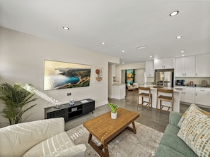 This modern living room features earth-toned decor, large windows for natural light, and cozy seating, including a swivel chair. A 65" TV is perfect for movie nights, while natural materials and soft lighting create a serene, inviting atmosphere. The space flows into the kitchen.