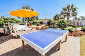 This vibrant backyard features a blue ping pong table shaded by a bright umbrella, offering a perfect spot for family fun amidst the lush landscaping and nearby mini-golf.
