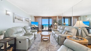 This spacious living room features plush reclining sofas, a large flat-screen TV, and stunning beach views through floor-to-ceiling glass doors—perfect for relaxing in comfort after a day in the sun.