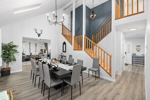 The dining room features a sleek table that comfortably seats 10 beneath a modern chandelier—ideal for hosting friends and family. It flows seamlessly into a warm and inviting living area, highlighted by a striking staircase set against a bold blue accent wall.