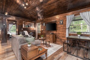 Relax in this rustic living room with rich wood tones, comfy seating, and a smart TV for cozy nights in 🪵📺. Large windows and warm lighting create a welcoming cabin vibe perfect for unwinding 🌲🛋️
