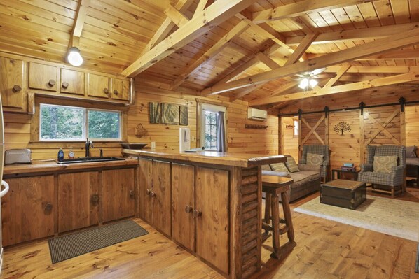 Cook and gather in this charming cabin kitchen with knotty wood cabinets, a peninsula bar with rustic stools, and vaulted beamed ceilings. Cozy living nook with plush seating and sliding barn doors adds to the rustic retreat vibe! 🪵🍳🔥