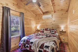 Cozy rustic bedroom with pinewood walls and vaulted ceiling. The bed features plaid bedding with bear and tree motifs, complemented by warm lighting and gray curtains framing a window with nature views 🌲🛏️🦌