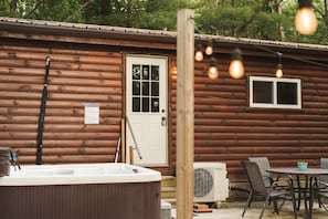 Cozy cabin exterior with inviting hot tub, patio seating area, and warm string lights—ideal for relaxing evenings in nature 🌟🛁🌲