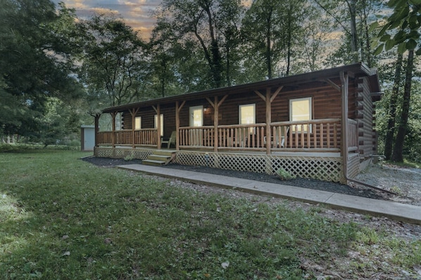 Rustic log cabin surrounded by lush trees, featuring a welcoming front porch with seating and warm glowing windows at dusk, perfect for a cozy retreat in nature 🌲🏡✨